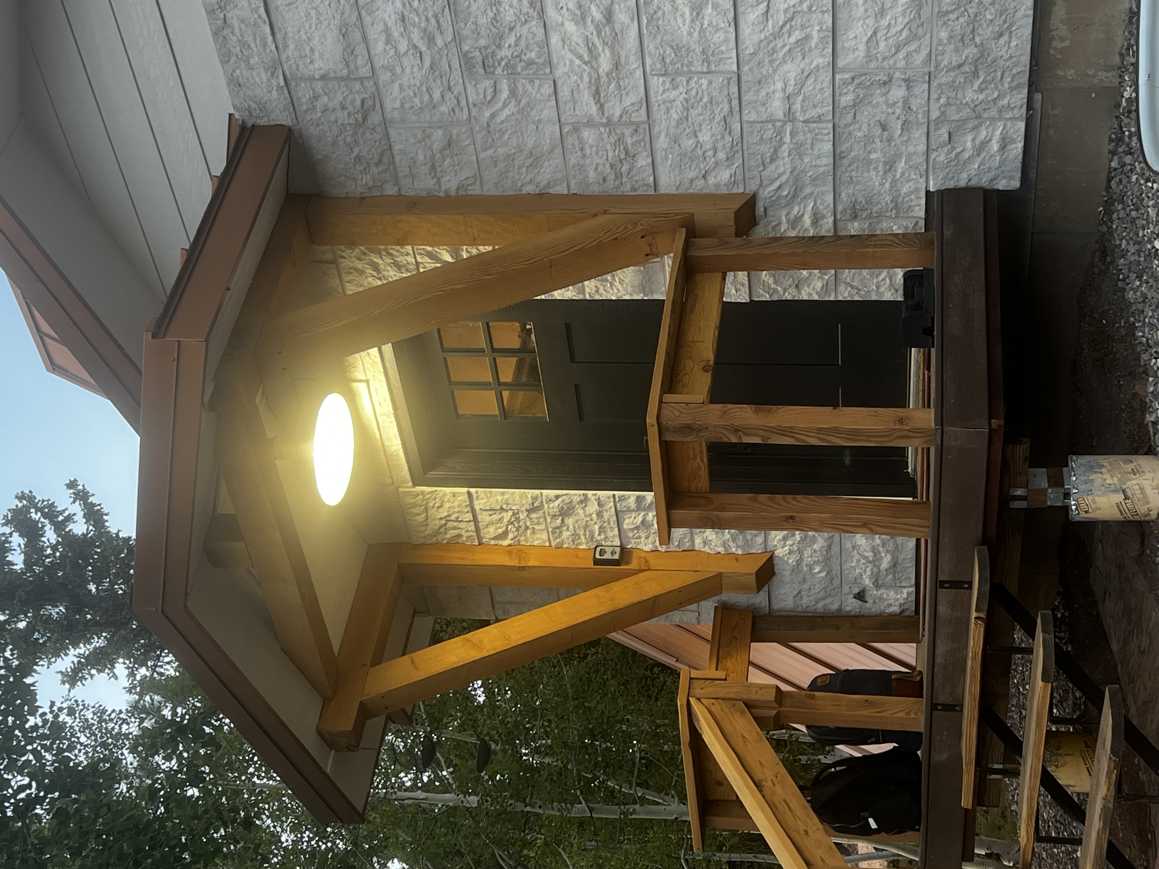 The front entry at dusk: timber-braced canopy lit by the porch light, stone veneer wall, black door, lockbox on the post.