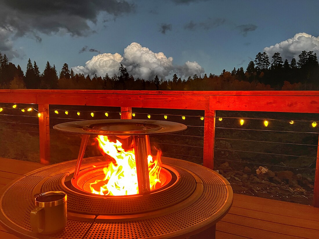 A pellet fire pit burning on the deck at dusk under string lights, with storm clouds over the treeline.