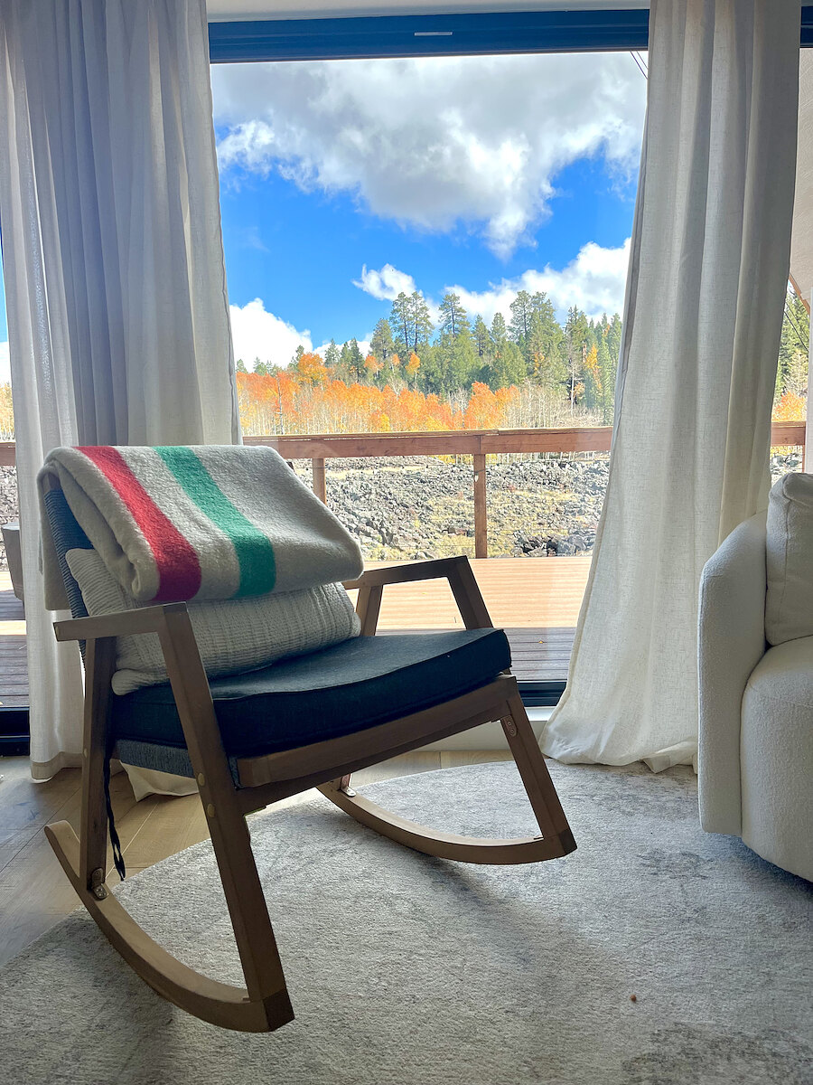 A rocking chair with a striped wool blanket beside the gable window, looking out at autumn aspens and the lava field.